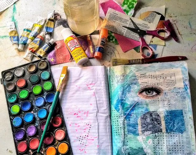 art journaling supplies: watercolor palette, paintbrush, notebook with collaged and painted images, acrylic paint tubes, water jar.